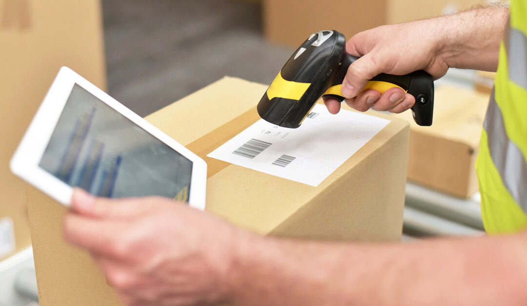 A worker holding a tablet in one hand and a scanning device in the other. They are scanning a barcode on a cardboard box.