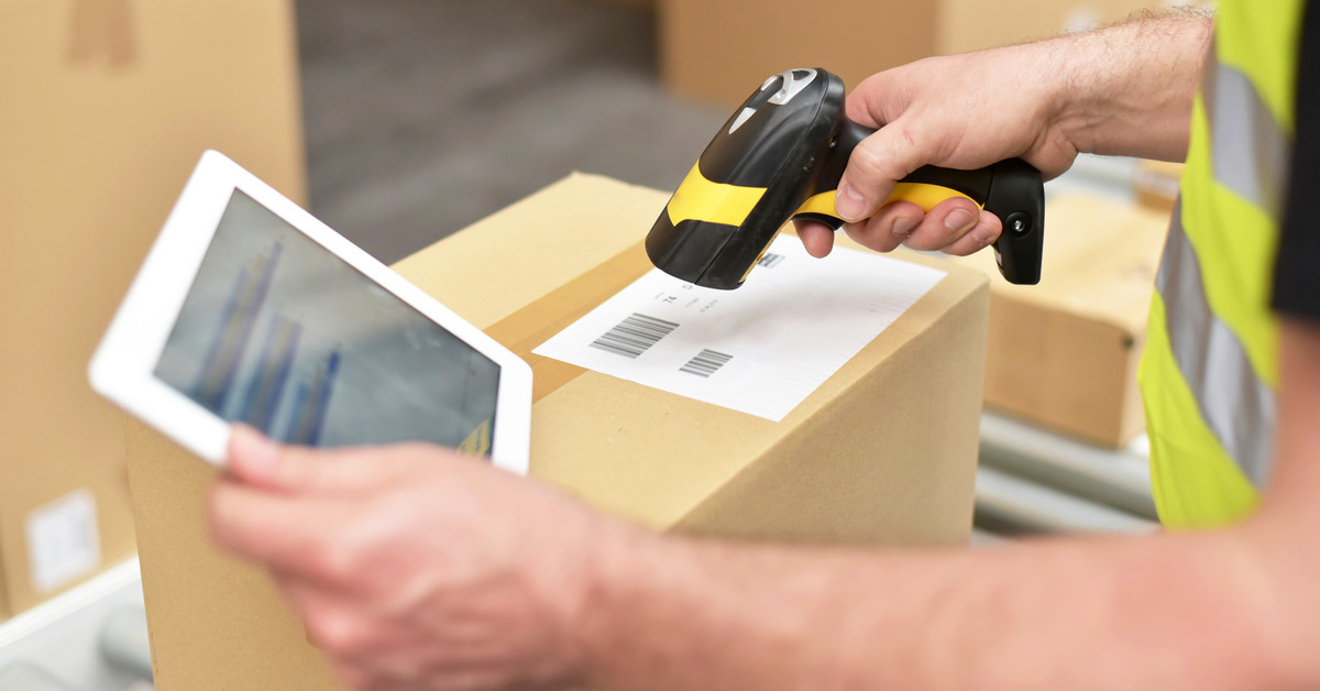 A worker holding a tablet in one hand and a scanning device in the other. They are scanning a barcode on a cardboard box.