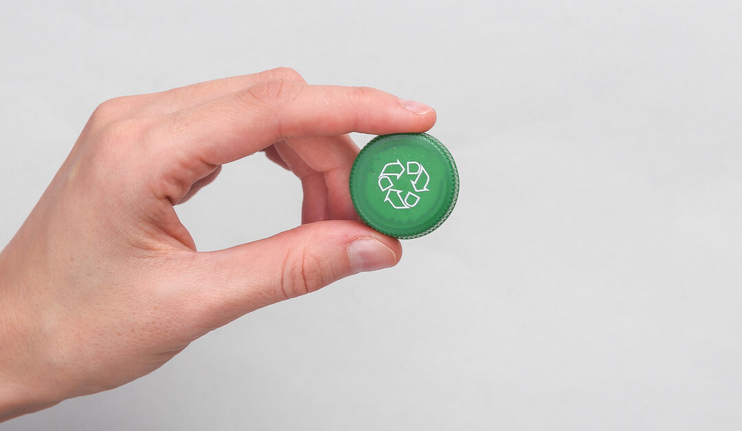 A hand holding a green plastic bottle cap against a white background. There is a recycle symbol on the plastic cap.