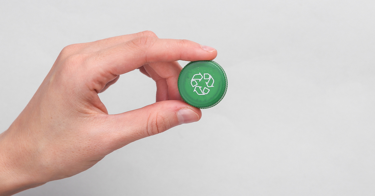 A hand holding a green plastic bottle cap against a white background. There is a recycle symbol on the plastic cap.