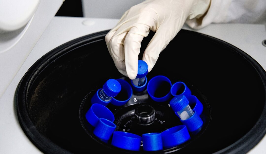 A medical device stores test tubes with blue plastic tube inserts. There is a gloved hand reaching for a test tube.