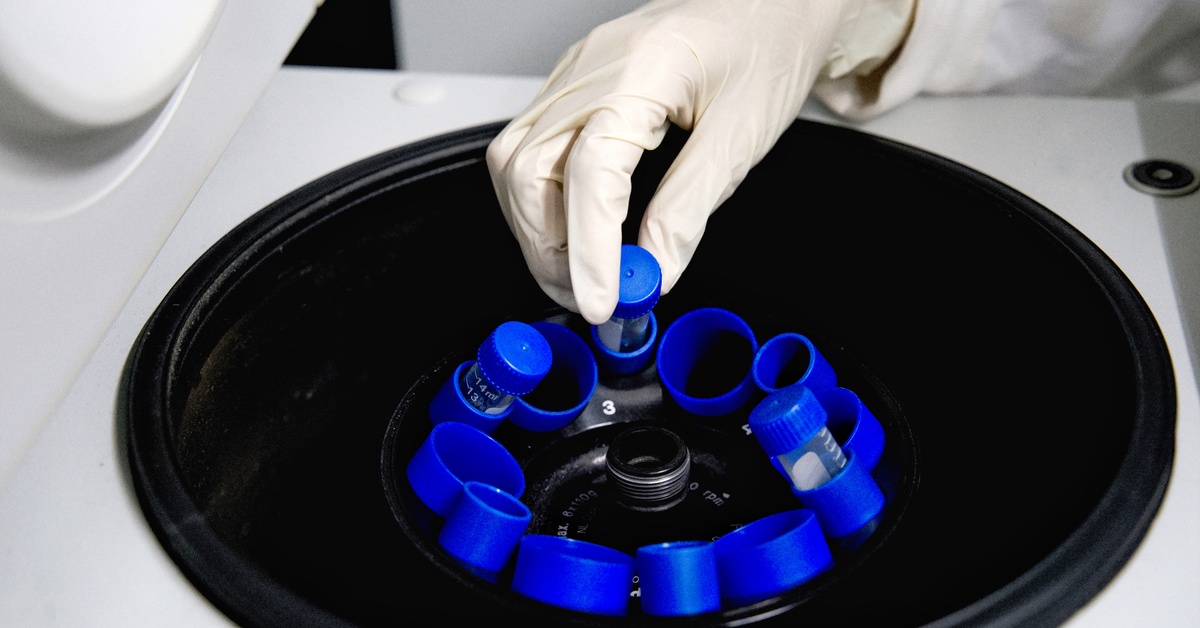 A medical device stores test tubes with blue plastic tube inserts. There is a gloved hand reaching for a test tube.