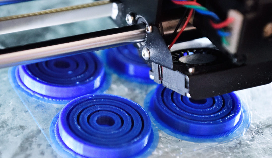 A 3D printing machine creating four blue round caps with ridges on the inside. The caps are sitting on a transparent surface.