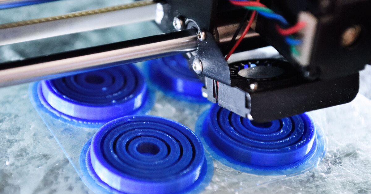 A 3D printing machine creating four blue round caps with ridges on the inside. The caps are sitting on a transparent surface.